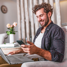 Smiling man relaxing at home while using his smartphone and laptop, showcasing how Adaptive Experiences create seamless, personalized digital interactions across multiple devices.